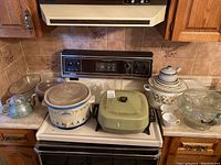 Overview of the entire lot with all items arranged on a stove and countertop, showing crock pot, griddle, plates, punch bowl, teapot, various glasses, and stock pot.