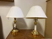 Pair of matching brass and white table lamps with white pleated fabric shades on a wooden surface against a wall.
