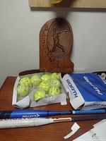 Wooden baseball wall hook, new packaged softballs and blue and white Worth box on wooden surface with two baseball bats placed below