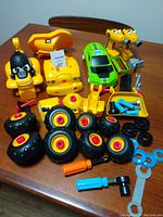 Full view of lot showing yellow CAT construction vehicle parts and wheels, green race car, yellow cartoon-style heads, and plastic tools on wooden surface.