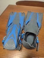 Pair of blue and gray snorkeling flippers with adjustable side straps, shown on wooden table.