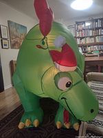 Front view of large green inflatable dinosaur with red spikes and Santa hat on its head, inflated and standing on a carpet indoors.