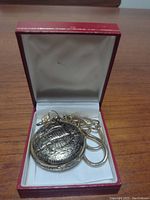 Closed view of the pocket watch in red lined box showing raised ship design on case.