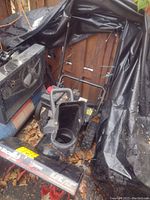 Front angle view of the snowblower showing the black red unit with chute, wheels, and handlebar, partially covered with a tarp, placed on leaves.