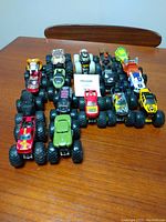 Top-down view of 14 assorted toy monster trucks placed on a wooden table showing various colors and designs with oversized wheels.