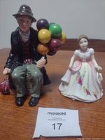 Two Royal Doulton porcelain figures placed on a wooden surface, showing Balloon Man holding bunch of balloons next to the Pretty Ladies figure in a floral dress.