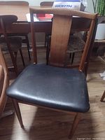 Side view of one teak wood chair showing black leather seat with minor tear and scuffed wood frame.