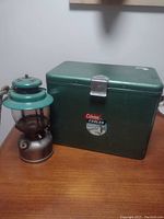 Full view of green Coleman cooler alongside green and silver Coleman lantern on wooden surface.