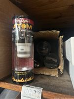 Photo of one First Alert Atom smoke and fire alarm in cylindrical packaging on shelf, showing front with logo and product features.