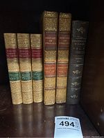 Photo showing six antique books standing vertically on a shelf with leather spines and gold embossed titles including 'Romans Under The Empire,' 'History of England,' and 'Thackeray's Works Vol 3'.
