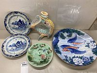 Shows front view of all five items including blue and white oval plates, green round plate with floral design, large round koi fish plate, and colorful fish-shaped vase.
