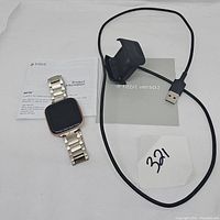 Fitbit Versa 2 watch with metal band alongside USB charger dock and instruction manuals on a white surface.