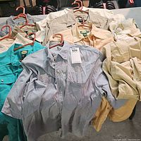 Top view photo showing all 11 men's short sleeve button-up shirts neatly hung on orange hangers. Colors include teal, various shades of khaki, beige, and blue. Each shirt has the Cabela's brand label and size 2XL tag visible on some.