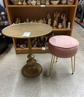 Photo of round matte gold metal table and pink velvet vanity stool with gold hairpin legs in front of wooden shelving.