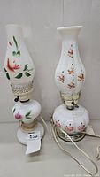 Pair of milk glass hurricane style lamps with floral painted designs, one with reddish-pink flowers and green leaves, the other with small pink flowers. Both show wear and have cords.