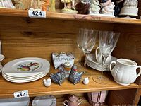 Wide shelf view showing stacks of decorative plates, salt and pepper shakers, clear wine glasses, small ceramic pitcher, bird figurines, and decorative box.