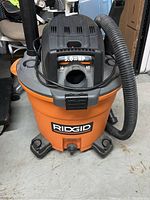 Front view of Ridgid wet dry vacuum showing orange tank, black top with power rating and hose attached.