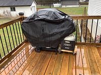 Covered grill with torn black protective cover on wooden deck outdoors in rain.