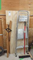 Full view of three plywood boards in different sizes, small metal two-step ladder, push broom.