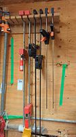 Full view of six bar clamps hanging on wooden wall rack with green tape marks indicating placement.