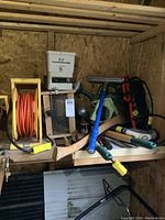 Photo showing the lot of garden tools on a wooden shelf, including extension cord on reel, bird feeder, hand seed spreader, bird house, axe, hose nozzle, and watering can.