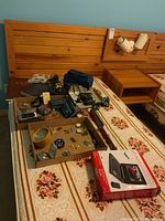 Wide view of lot showing assorted electronics, collectibles, and books on bed with wooden headboard and tables in background