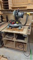 Wide view showing Delta mitre saw mounted on custom wooden table with wheels, shelves holding saw blades, outlet and switch on table side.