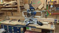 Wide angle photo showing the Kobalt 10" mitre saw mounted on a wooden table with blue organizer boxes underneath. Includes multiple metal rollers on the table edges.