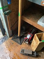 Tools arranged together near wooden shelving, showing green-headed sledgehammer, sand stamper, red hydraulic car jack, metal scissor jack, and wooden box.