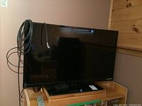 Image showing front view of Dynex 40 inch TV placed on wooden stand. Several cables are hanging on left side.