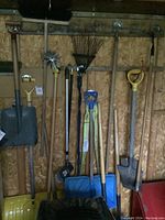 Wide shot of yard tools hanging on wooden wall, shows shovels, hoes, rakes, hand tiller, broom, and blue snow shovel