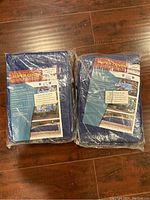 Photo showing two unopened packs of blue tarps on a wooden floor. Each pack contains two tarps, making four tarps total. Packaging label 'Super Cover' visible, indicating size 8x10 feet.