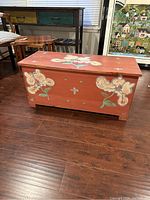 Front and side view of the antique red stained wooden trunk featuring hand-painted white and blue flowers on the front panel and top surface, with visible cross and dot accents.
