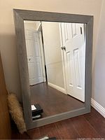Full view of the wood framed mirror leaning against a wall, showing the reflective glass and the grey painted frame with visible wood grain.