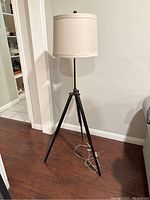 Full view of the metal tripod base floor lamp with cream cylindrical shade, placed on a wooden floor against a white wall