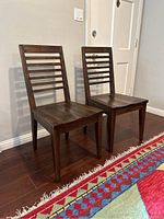 Two Riverside Furniture dining chairs with slat-back design, showing front and side angles on hardwood floor with colorful rug in foreground.