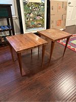 Two wooden square side tables with tapered legs shown standing on dark wood floor near framed art and other furniture. Tables show worn wood surface with scratches and discoloration.