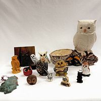Photo of 14 owl figurines arranged on a white background, showing variety in size, shape, color, and material. Largest piece is white ceramic owl with bowl.