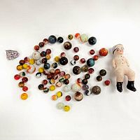 Top-down image showing assortment of various sized glass marbles, small seated painted clay figure, and a small purple stone arranged on a white surface.