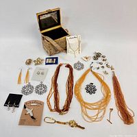 Photo displaying fabric-covered box, jewelry items like beaded necklaces, metal snowflakes, pins, earrings, and a watch arranged on white surface.