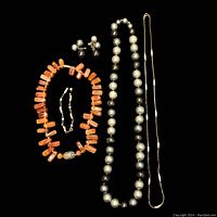 Top view of the jewelry lot on black background showing red-orange bead necklace and bracelet, silver and black pearl necklace, delicate chain necklace, and matching ear clips.