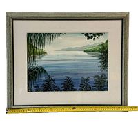 Framed watercolor painting showing a tranquil water scene with plants in foreground and mountains in background, measuring approximately 21.5 inches wide as shown by tape measure placed below frame.