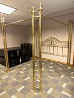 View of large polished metal canopy bed frame showing the tall posts with rounded finials and a horizontal canopy bar at the top. The polished metal surface is reflective, and reinforcement bars are noted as included but not visible.