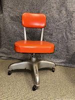 Frontal view of the vintage low metal rolling chair showing orange vinyl padded seat and backrest, five metal legs with caster wheels.