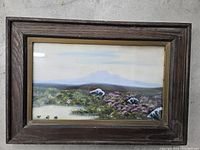 Framed landscape painting showing the entire composition including rocks, trees, and hills, behind glass in a dark wooden frame.