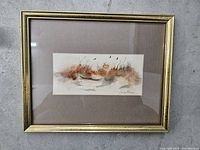 Framed watercolor artwork showing muted earth tone grasses and birds under glass in a gold frame.
