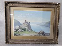 Full view of framed vintage artwork depicting a castle on a rocky hill by a lake with mountains in the background.