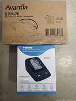Photo of both blood pressure monitors' original packaging. Avantia BPM-70 in brown box on top of a blue and white Vimmik DBP-6191 box showing monitor image and features.