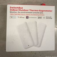 Front of SwitchBot Indoor/Outdoor Thermo-Hygrometer box showing product name, device image, and logos of compatible smart home systems.