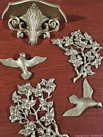 Five silver-colored metal wall decorations on a wooden floor: an ornamental leaf and scroll, two birds in flight, and two floral branches.